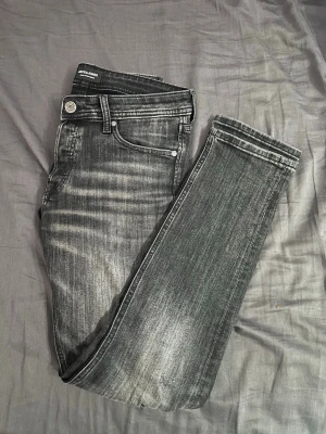 Grå slim fit jeans från Jack & Jones - Säljer ett par grå slim fit jeans från Jack & Jones med snyggt slitna detaljer och klassisk femficksdesign. Jeansen har en silverfärgad knapp och dragkedja, samt tydliga kontrastsömmar. Materialet är bomullsdenim med stretch för extra komfort. Som man kan se finns det ett hål vid knät men de e är ett lättfix och synns inte särskillt mycket igentligen