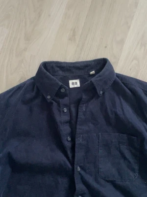 Mörkblå Manchester overshirt från Uniqlo - Stilren mörkblå overshirt från Uniqlo med klassisk button-down krage och bröstficka. Tillverkad i ett mjukt, lite tjockare Manchester tyg som passar perfekt som lager. Långa ärmar och tidlös design gör den enkel att matcha till olika outfits.