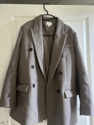 Grå dubbelknäppt kavaj - Snygg grå kavaj med dubbelknäppning och breda slag. Kavajen har två fickor med lock och en bröstficka. Tillverkad i ett mjukt, ull-liknande material och helfodrad för extra komfort. Perfekt för en stilren och trendig look.