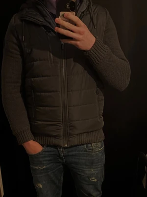 Mörkgrå dunväst med stickade ärmar - Snygg mörkgrå dunväst med dragkedja framtill och två sidofickor. Västen har stickade ärmar och ribbade muddar för extra komfort och stil. Perfekt för lager-på-lager och ger en modern look till din outfit.