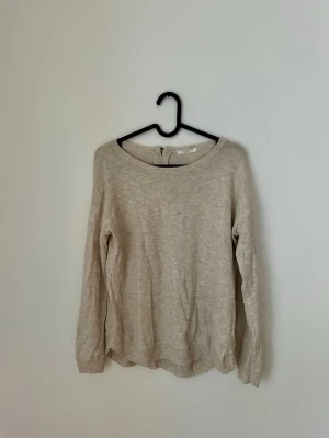 Beige stickad tröja med dragkedja - Beige stickad tröja med rund halsringning och dekorativ dragkedja bak i nacken. Tröjan har långa ärmar och ribbade muddar vid ärmslut och nederkant. 