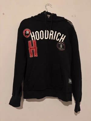 Hood Rich tracksuit  - Snygg svart hoodie från Hoodrich med stora vita och röda broderade detaljer och patches på bröstet. Klassisk huva med dragsko och stor magficka framtill. Perfekt för dig som gillar streetwear och vill sticka ut lite extra.