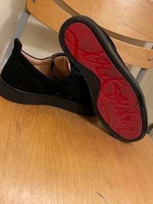 Svarta Louboutin skor - Snygga svarta sneakers från Christian Louboutin i mocka med klassisk röd sula och svart snörning. Låg modell med diskret design och exklusiv känsla. Perfekta för dig som vill sticka ut med en ikonisk detalj under skon.