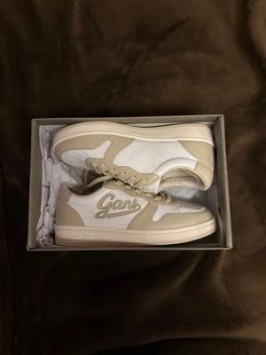 Beige och vita sneakers från Gant - Snygga sneakers från Gant i beige och vitt med broderad logga på sidan. Skorna har en klassisk låg siluett, vita snören och detaljer i mocka och läder. Perfekta för en clean och stilren look. Kommer i originalkartong.