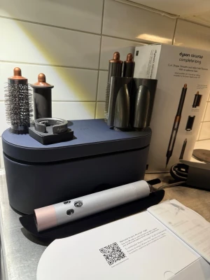 Dyson Airwrap Complete Long hårstyler - Säljer en Dyson Airwrap Complete Long med flera stylingtillbehör. Setet innehåller olika borstar och cylindrar för att skapa lockar, volym och släta frisyrer. Färgen är silver med kopparfärgade detaljer och allt kommer i ett snyggt mörkblått fodral.