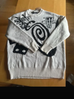 Vit stickad tröja från stüssy - Säljer en vit stickad tröja med svart grafiskt mönster både fram och bak. Tröjan har rund halsringning, ribbade muddar och nederkant. Mönstret består av spiraler, symboler och en åtta-boll vilket ger en unik och modern look. Taggen sitter kvar