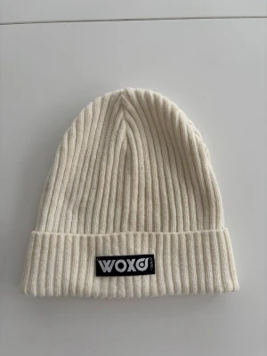 Vit ribbad mössa från WOXO - Säljer en stilren vit ribbstickad mössa från WOXO. Mössan har uppvikt kant och en svart patch med WOXO-loggan framtill. Tillverkad i mjukt och stretchigt material som sitter skönt på huvudet. Perfekt för kalla dagar och enkel att matcha med olika outfits.