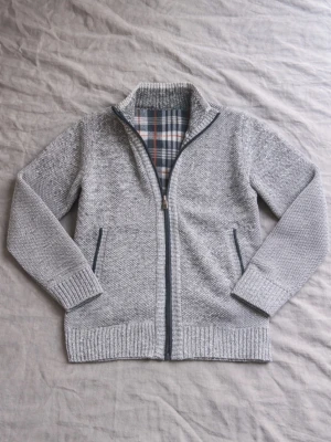 Grå stickad cardigan med dragkedja - Snygg grå stickad cardigan med dragkedja och ribbade muddar. Insidan har ett rutigt foder i blått, vitt och orange. Två fickor framtill med mörkare kant. Perfekt för lager-på-lager och chill stil.