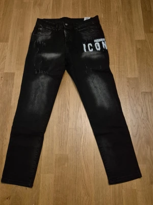 Svarta Dsquared2 ICON jeans - Säljer ett par svarta jeans från Dsquared2 med vit 'ICON'-text på ena sidan framtill. Jeansen har en slim fit passform, slitna detaljer och klassisk femficksdesign. Materialet är denim med stretch för extra komfort. Perfekta för en trendig streetwear-look.