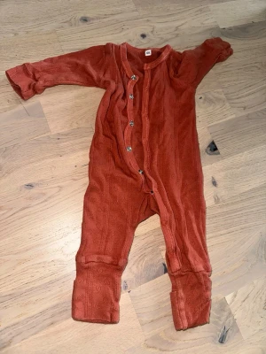 Rostfärgad babybody med knappar - Superfin långärmad babybody i rostfärg med dekorativa knappar framtill. Bodyn har ett diskret hålmönster i tyget och är tillverkad i mjukt material som känns skönt mot huden. Perfekt för mysiga dagar hemma.