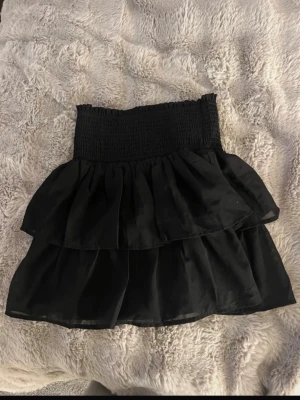 Svart volangkjol från Bubbleroom - Supersöt svart kjol från Bubbleroom i storlek XS. Kjolen har smockad midja och två lager volanger som ger en snygg och trendig look. Tillverkad i ett lätt och luftigt tyg som känns bekvämt hela dagen.