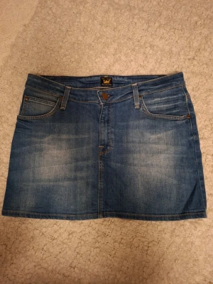 Blå jeanskjol från Lee - Säljer en klassisk blå jeanskjol från Lee med fem fickor och bälteshällor. Kjolen har en kort modell och är tillverkad i slitstarkt denimtyg. Perfekt att matcha med sneakers eller boots för en avslappnad look. W29