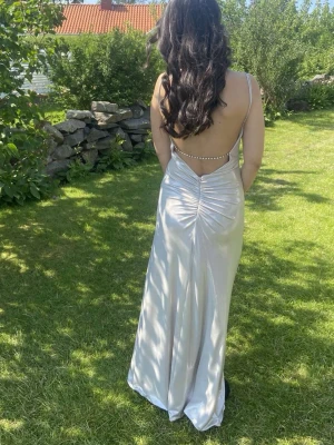 Silverfärgad lång klänning med öppen rygg - Säljer en elegant silverfärgad långklänning med tunna axelband och öppen rygg dekorerad med pärldetaljer. Klänningen har en figurnära passform och skimrande material som ger en lyxig känsla. Perfekt för bal eller festkväll.
