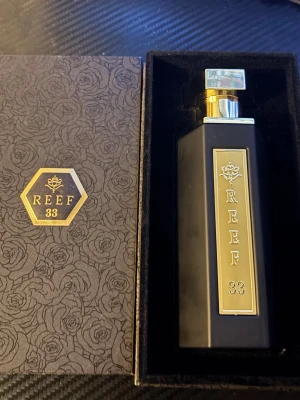 Reef 33 exklusiv parfymflaska - Snygg parfym från Reef med elegant svart och guld design. Flaskan är rektangulär med guldfärgad metallplatta och detaljer, samt genomskinlig fyrkantig kork. Kommer i en lyxig svart ask med rosor och guldfärgad logotyp. Perfekt för dig som gillar exklusiva dofter.