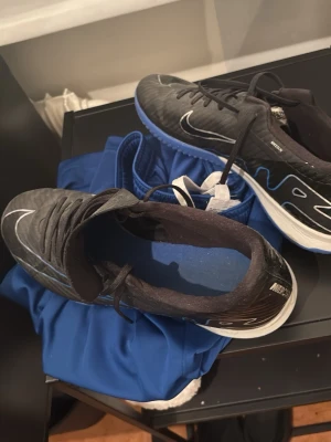 Nike Mercurial Air Zoom fotbollsskor - Säljer ett par Nike Mercurial Air Zoom fotbollsskor i svart med blå och vita detaljer. Skorna har snörning, låg profil och en blå sula med greppmönster, perfekt för spel på konstgräs eller inomhus. Tillverkade i syntetmaterial för lätt känsla och bra bollkontroll.