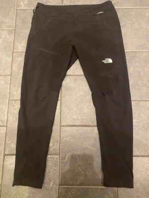 Svarta vindbyxor The North Face - Säljer ett par svarta vindbyxor från The North Face med smal passform och dragkedjeficka på sidan. Byxorna har en diskret logga på låret och är tillverkade i ett mjukt och stretchigt material som känns skönt mot huden.