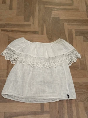 Vit offshoulder blus med spets - Superfin vit offshoulder blus med bred spetsdetalj runt axlarna. Luftigt material som passar perfekt till varma dagar. Snygg och enkel att matcha med både jeans och kjol.