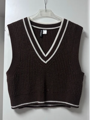 Brun stickad väst från H&M - Snygg brun stickad väst från Divided med vita ränder vid v-ringad hals och nederkant. Croppad modell i ribbstickat material, perfekt att styla över en skjorta eller t-shirt. Klassisk och trendig look.