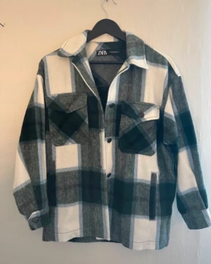 Rutigt overshirt från Zara - Snygg overshirt från Zara med stora rutmönster i mörkgrönt, grått och vitt. Modellen har två bröstfickor med knappar, klassisk krage och knäppning framtill. Perfekt att slänga över en t-shirt för en avslappnad look.