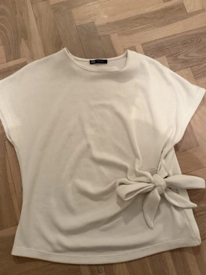 Vit topp med knyt från Zara - Stilren vit topp från Zara med korta ärmar och rund halsringning. Toppen har en snygg knytdetalj i sidan som ger en extra touch. 