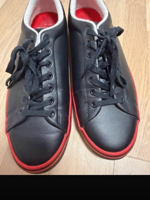 Svarta Polo sneakers med röd sula - Snygga svarta sneakers från Polo med röd sula och röd insida. Skorna har svarta skosnören, lågt skaft och Polo-logga bak på hälen. Ovandelen är i slätt material som liknar läder och insidan är mjukt fodrad i vitt och rött. Helt nya 