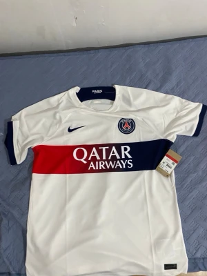 PSG vit matchtröja Nike - Snygg vit matchtröja från Paris Saint-Germain med blå och röd rand över bröstet. PSG-logga och Nike Swoosh på bröstet samt Qatar Airways tryckt framtill. Tillverkad i lätt och ventilerande Dri-FIT material, perfekt för fotboll eller träning.