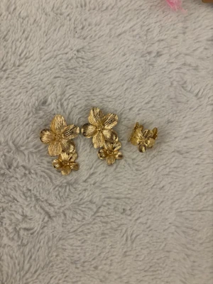 Blom örhängen med matchande ring - Unika örhängen och ring i guldfärgad metall med detaljerade blommor i lager på lager-design. Blommorna har en elegant, strukturerad yta som ger ett exklusivt intryck. Perfekta för dig som vill sticka ut med ett trendigt accessoarval. Köpt på marknad i Spanien 