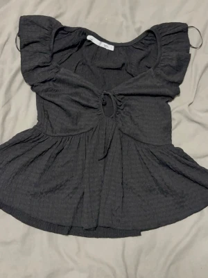 Svart topp med knyt från 157 - Supersnygg svart topp från 157 med korta ärmar och knyt framtill. Toppen har en lätt rynkad struktur och en flowy passform nedtill. Perfekt att styla med jeans eller kjol för en trendig look.