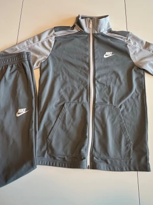 Nike grå träningsset med dragkedja - Nike träningsset i grått med vita detaljer. Jackan har hel dragkedja, ståkrage och två fickor framtill. Byxorna har resår i midjan och Nike-logga på benet. Perfekt för träning eller chill. Materialet är mjukt och följsamt, troligen polyester.
