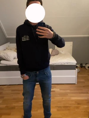 Mörkblå hoodie från Les Deux - Snygg mörkblå hoodie från Les Deux med vit logga på bröstet. Klassisk modell med huva och dragsnören samt en stor magficka. Tillverkad i mjukt material som passar perfekt till jeans eller joggers. Enkel och stilren design som funkar till det mesta. Passar dig mellan 170-180 cm