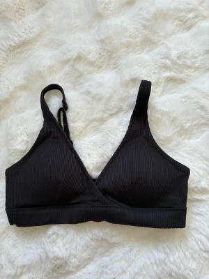 Svart ribbad bikiniöverdel - Snygg svart bikiniöverdel med ribbad struktur och justerbara axelband. Modellen har omlott-skärning framtill och stängs med spänne i ryggen. Vet ej storlek, skulle gissa runt 34 eller Xs!