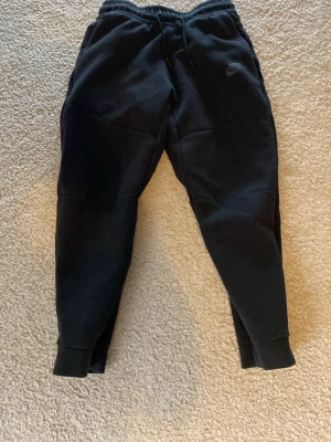 Svarta Nike mjukisbyxor - Svarta mjukisbyxor från Nike med dragsko i midjan och ribbade muddar vid benslut. Byxorna har en avslappnad passform och är tillverkade i mjukt material, perfekt för chill eller träning. Diskret Nike-logga på låret.