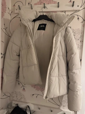 Ljusbeige pufferjacka från ZARA - Säljer en ljusbeige pufferjacka från ZARA med hög krage och stor huva. Jackan har quiltad design, dragkedja framtill och är tillverkad i ett mjukt, vadderat material som håller värmen. Perfekt för kalla vinterdagar och riktigt snygg till alla outfits.💗