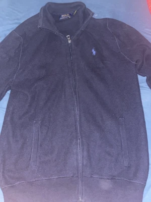 Mörkblå cardigan från Polo Ralph Lauren - Snygg mörkblå cardigan från Polo Ralph Lauren med hög krage och dragkedja framtill. Tröjan har två fickor och den klassiska broderade loggan på bröstet. Perfekt till lager på lager och enkel att matcha med jeans eller chinos. Inga skador eller hål. Priset går och pruta!