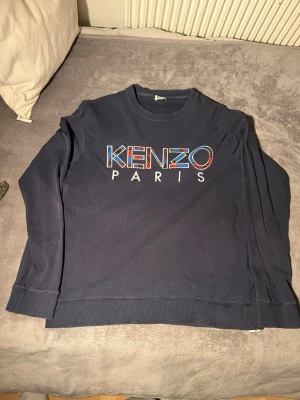 Mörkblå Kenzo sweatshirt - Snygg mörkblå sweatshirt från Kenzo med stort broderat Kenzo Paris-tryck i färgglada detaljer på bröstet. Tröjan har rund halsringning, ribbade muddar och är tillverkad i mjukt bomullsmaterial. Perfekt för en trendig och avslappnad stil.