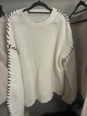 Stickad tröja  - Säljer en oversized vit stickad tröja med svarta tvinnade detaljer längs axlar och ärmar. Tröjan har rund halsringning, långa ärmar och ribbade muddar. Perfekt för dig som gillar en enkel men ändå unik look.