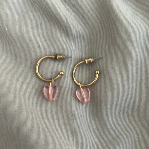 Guldiga örhängen med rosa hjärtan - Superfina örhängen i guldfärgad metall med hängande rosa hjärtan! Perfekt för dig som vill ha en söt och lekfull detalj till din look. Kommer aldrig till användning längre. OBS, katt finns i hemmet!