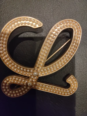 Guldfärgad brosch med pärlor - Snygg brosch i form av bokstaven L, guldfärgad metall dekorerad med små vita pärlor över hela ytan. Perfekt accessoar för att ge din outfit en personlig touch. Broschnålen sitter på baksidan för enkel fastsättning.