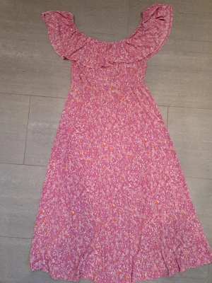 Rosa blommig långklänning volang - Söt rosa långklänning med blommigt mönster och bred volang runt axlarna. Klänningen har en offshoulder-design och är tillverkad i ett lätt, mjukt tyg som passar perfekt till varma dagar. Färgerna går i rosa, vitt och lite orange.