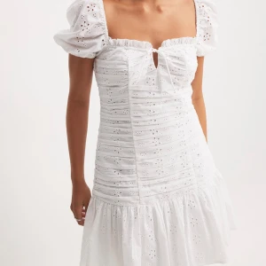 Vit kort klänning - Superfin vit kort klänning med puffärmar, broderade detaljer och volang nedtill. Klänningen har fyrkantig urringning, liten rosett framtill och är tillverkad i bomull. Perfekt för sommarens alla tillfällen och ger en romantisk vibe. Helt oanvänd med prislapp kvar och helt slutsåld på NAKD