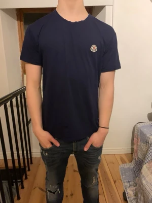 Mörkblå Moncler t-shirt - Säljer en mörkblå t-shirt från Moncler med klassisk passform och rund hals. T-shirten har korta ärmar och en liten Moncler-logga på bröstet. Perfekt till jeans eller shorts för en clean och enkel stil.