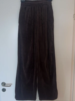 Bruna vida mjukisbyxor i velour - Mörkbruna mjukisbyxor med breda ben och resår i midjan. Tillverkade i mjuk velour som ger en skön och avslappnad känsla. Perfekta för en chill och trendig look. Har 2 sidofickor 