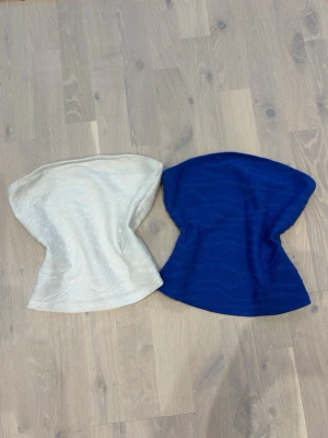 Vita och blå bandeau tubtoppar - Två bandeau tubtoppar i vitt och blått med diskret strukturmönster. Toppmodellen är axelbandslös och har en lätt utsvängd form nertill. Perfekta att styla till jeans eller kjol för en trendig look.