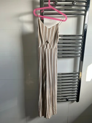 Beige randig jumpsuit med cutout - Säljer en beige jumpsuit med vita vertikala ränder och smala axelband. Den har en snygg cutout-detalj framtill och vida ben. Perfekt för dig som gillar en avslappnad och trendig stil. Materialet känns lätt och luftigt. I storlek 32 💞