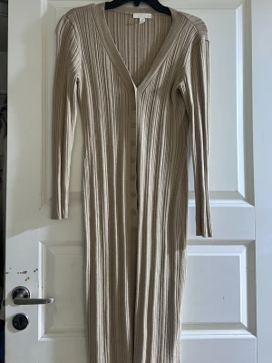 Beige ribbad långklänning med knappar - Säljer en beige ribbad långklänning med v-ringning och långa ärmar. Klänningen har knappar hela vägen framtill och är i ett mjukt stickat material som sitter snyggt på kroppen. Perfekt för dig som gillar stilrena och bekväma plagg.