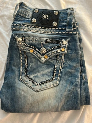 Miss Me blå jeans med dekorerade fickor - Säljer ett par blå Miss Me skinny jeans med slitningar och dekorativa bakfickor. Fickorna har broderade detaljer, nitar och glittriga stenar. Jeansen har normal midja och raka ben. Materialet är klassisk denim i bomull med snygg tvättad look.