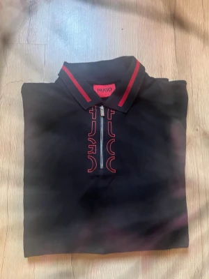 Svart Hugo Boss skjorta med dragkedja - Svart t-shirt från Hugo Boss med röda detaljer och dragkedja framtill. Klassisk krage med röda ränder och tryckt Hugo-logga längs dragkedjan. Regular fit och modern look, perfekt för dig som gillar stilrena plagg med en twist.