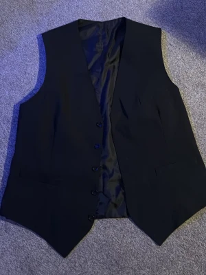 Svart kostymväst med knappar - Stilren svart kostymväst med klassisk passform och fem knappar framtill. Västen har slät yta och diskreta fickor på bröstet