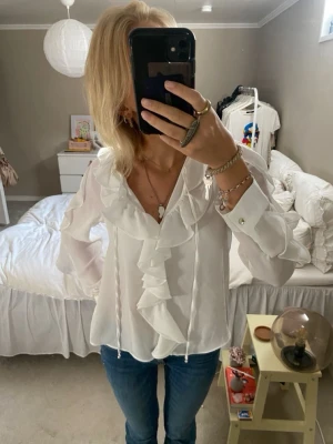 Vit blus med volanger och knytband - Säljer en snygg vit blus med volanger längs v-ringningen och knytband framtill. Blusen har långa ärmar med manschett och knappdetalj. Perfekt att matcha med jeans för en trendig look.