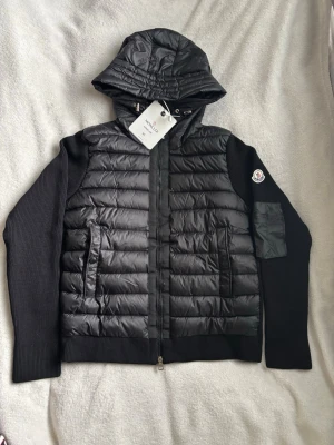 Svart pufferjacka från Moncler - Snygg svart pufferjacka från Moncler med stickade ärmar och huva. Jackan har quiltad front, dragkedja, två fickor och Moncler-logga på ärmen. Bröst:51cm längd: 61cm arm:61cm